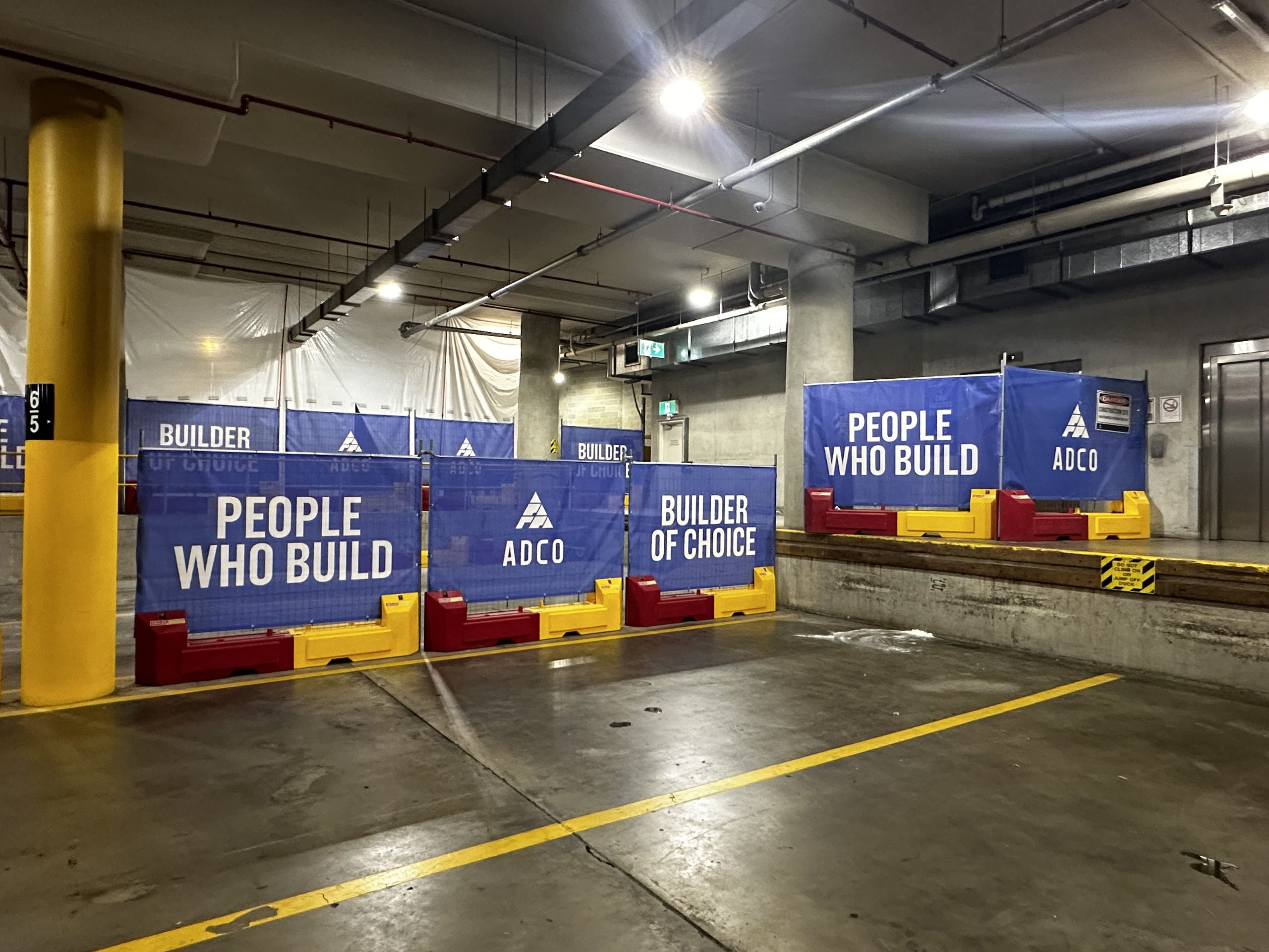 Temporary defence fencing, blue and yellow barriers with People Who Build and Builder of Choice displayed in an indoor parking area.