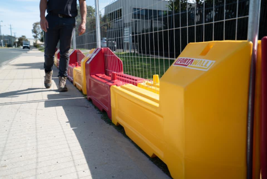 Gallery: FortaWall safety barriers in red and yellow, lining a sidewalk next to a chain-link fence. A person walks past the barriers.