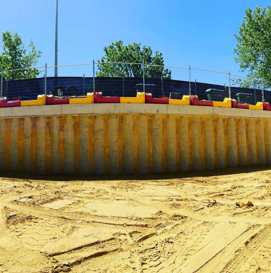 Gallery: Retaining wall with yellow & red safety barriers, chain-link fence, & sand. Tire tracks in the sand. Trees & clear blue sky in the background.