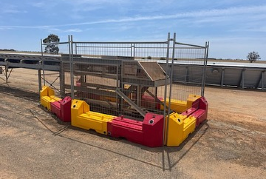 Gallery: A metal structure with yellow and red plastic barriers sits on a dirt surface. A conveyor belt and a long, low building are visible in the background.