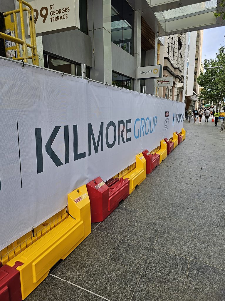 Gallery: Kilmore Group construction site barrier on a city sidewalk. Red and yellow plastic barriers line the bottom. Building visible in background.