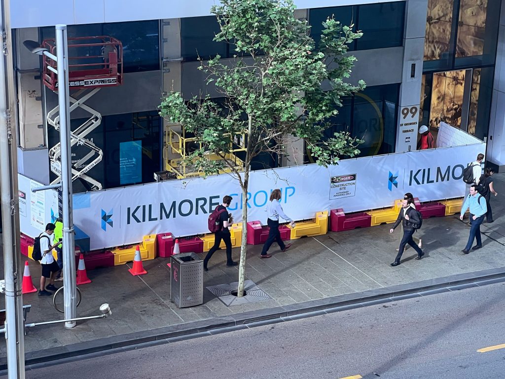 Gallery: Kilmore Group construction site on a city street. People walk along the sidewalk in front of the barrier. Construction equipment is visible.