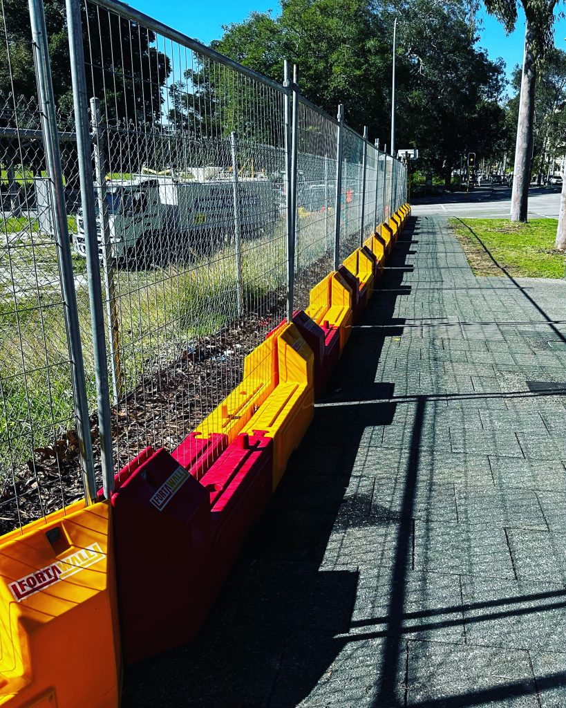 Gallery: Metal fence with yellow and red plastic barriers along a sidewalk. The fence is likely for construction or crowd control.