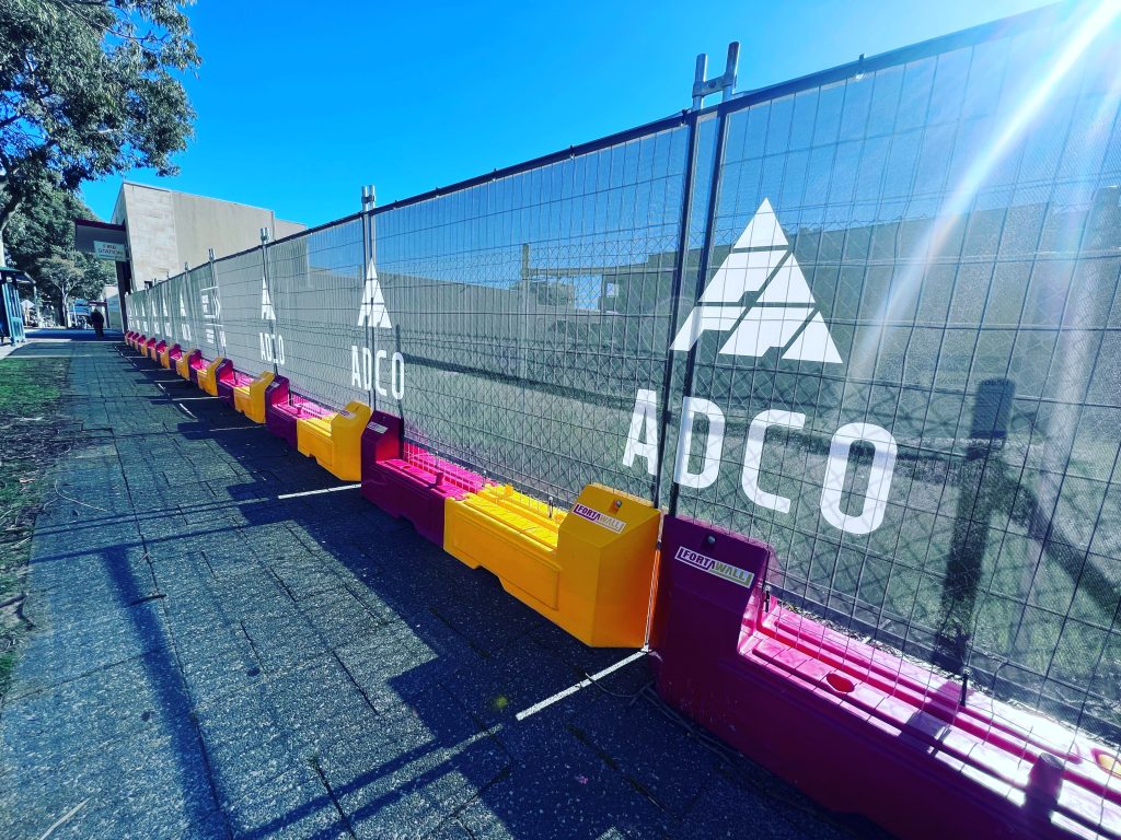 Gallery: Construction site barrier with ADCO logo. Yellow and red plastic bases support a wire mesh fence on a sunny day. Sidewalk visible.