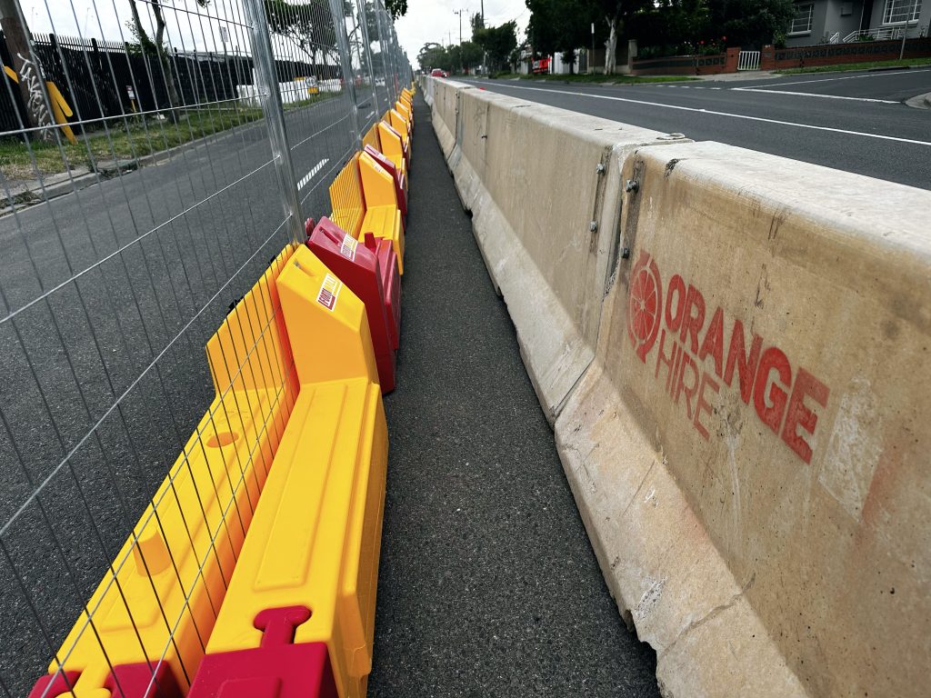 Gallery: Road safety barriers, including concrete and yellow/red plastic, line a street. Orange Hire is printed on the concrete barrier.