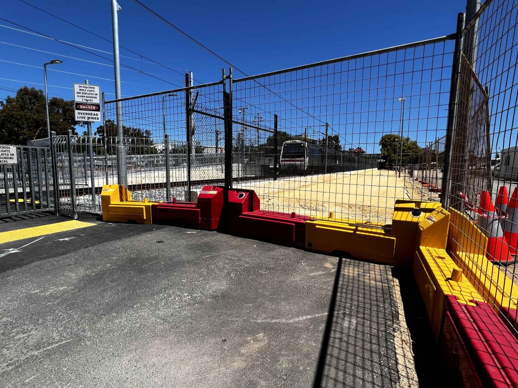 Gallery: Train station construction site with safety barriers, fencing, and warning signs under a clear blue sky. Train visible in background.