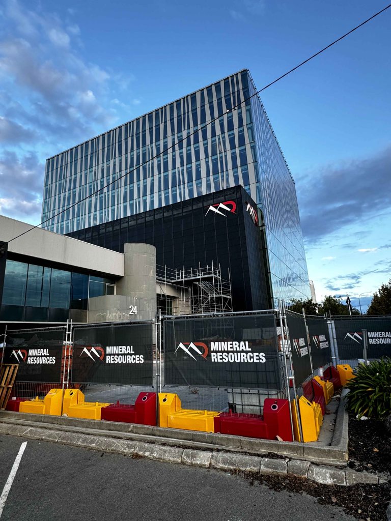 Gallery: Mineral Resources building with a modern glass facade. Construction barriers are in place, featuring the company's logo. Cloudy sky above.