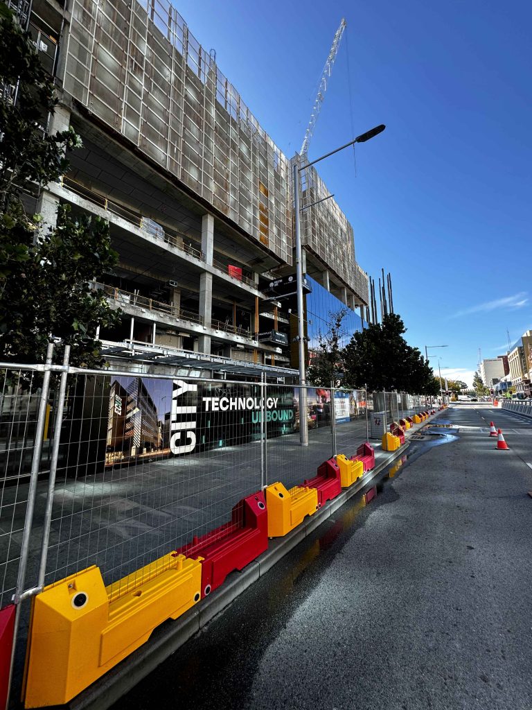 Gallery: Construction site with unfinished building, crane, and barriers. City Technology Unbound banner on the fence. Street view on a sunny day.