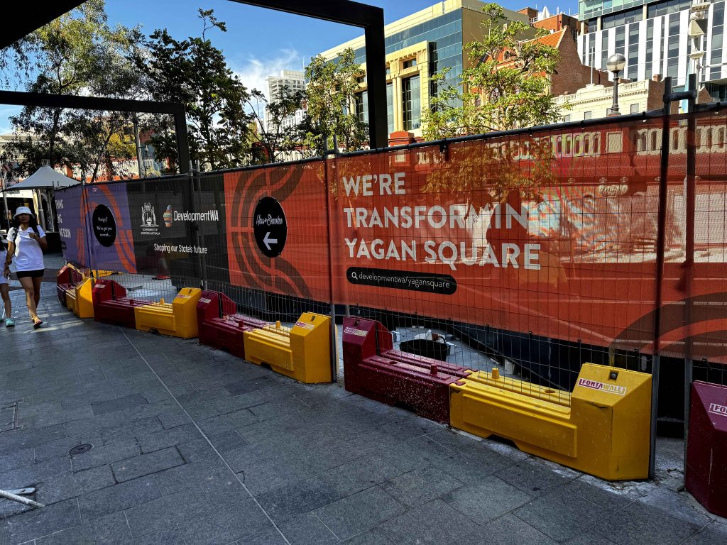 Gallery: Construction at Yagan Square. Fencing with We're Transforming Yagan Square banner. Yellow & red barriers line the perimeter. Pedestrians walk by.