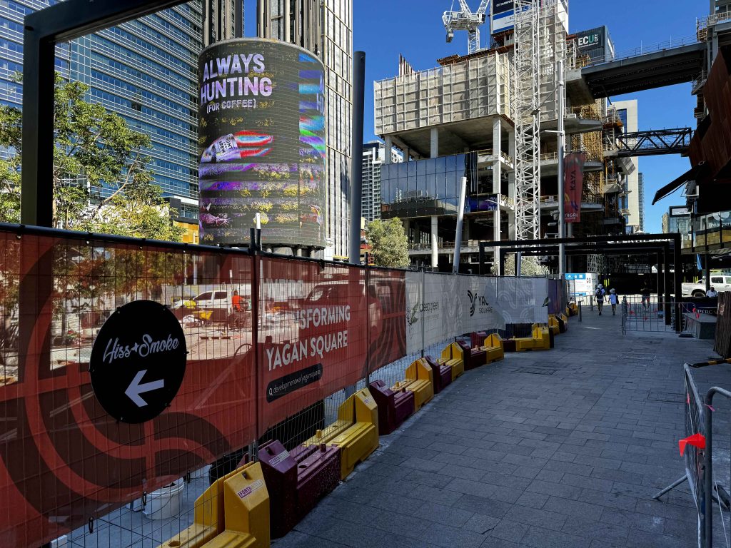 Gallery: Yagan Square construction site with fencing, signage, and a digital billboard. Tall buildings and a crane are visible in the background.