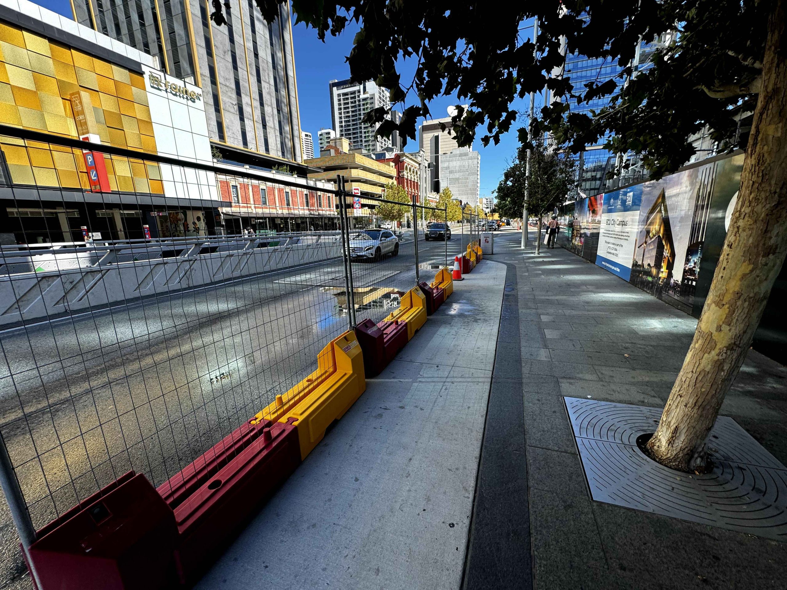 Gallery: City street view with barriers, buildings, and a tree. Construction site visible with fencing and posters. Cars on the road. Sunny day.