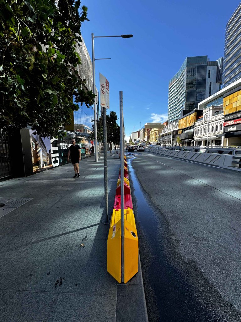City street view from Gallery. Yellow & red barriers divide the road. Buildings & shops line the street, with a clear blue sky above. Pedestrian on the left sidewalk.