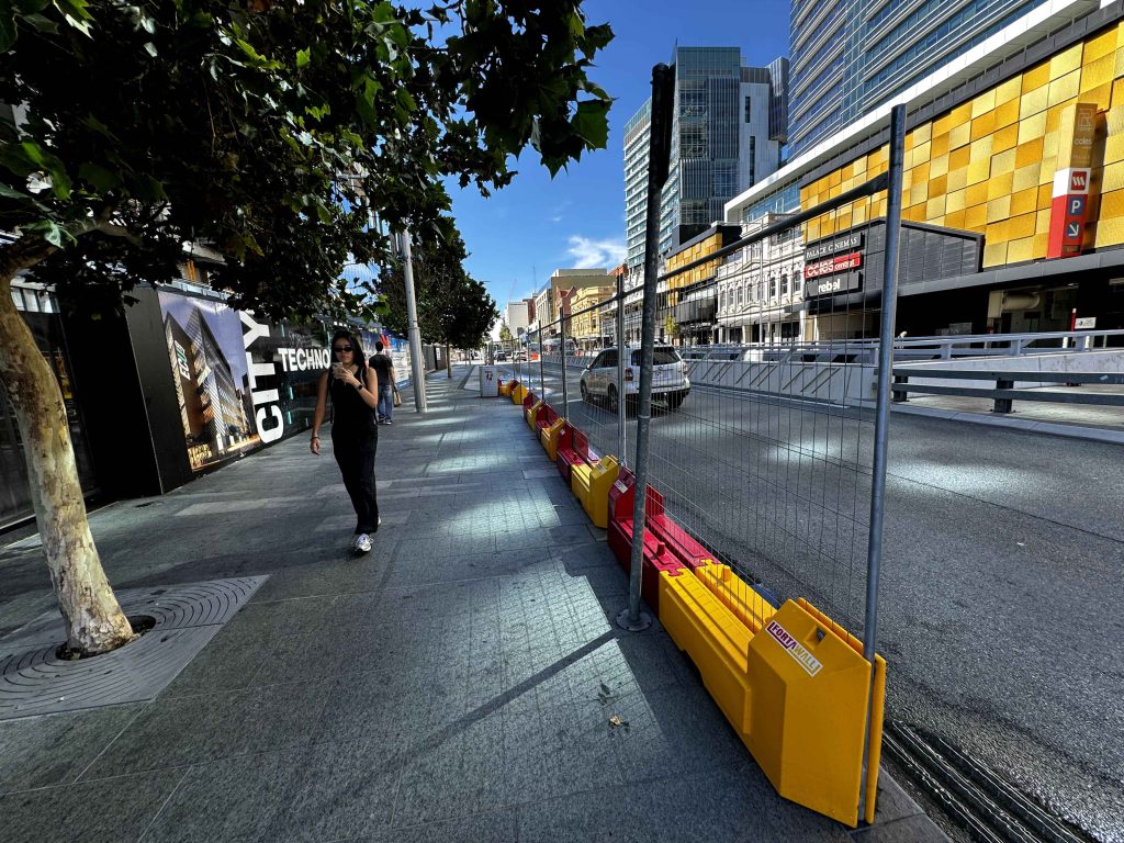 Gallery: City street view with barriers, buildings, and pedestrians. A woman walks on the sidewalk past construction hoarding. Modern architecture.