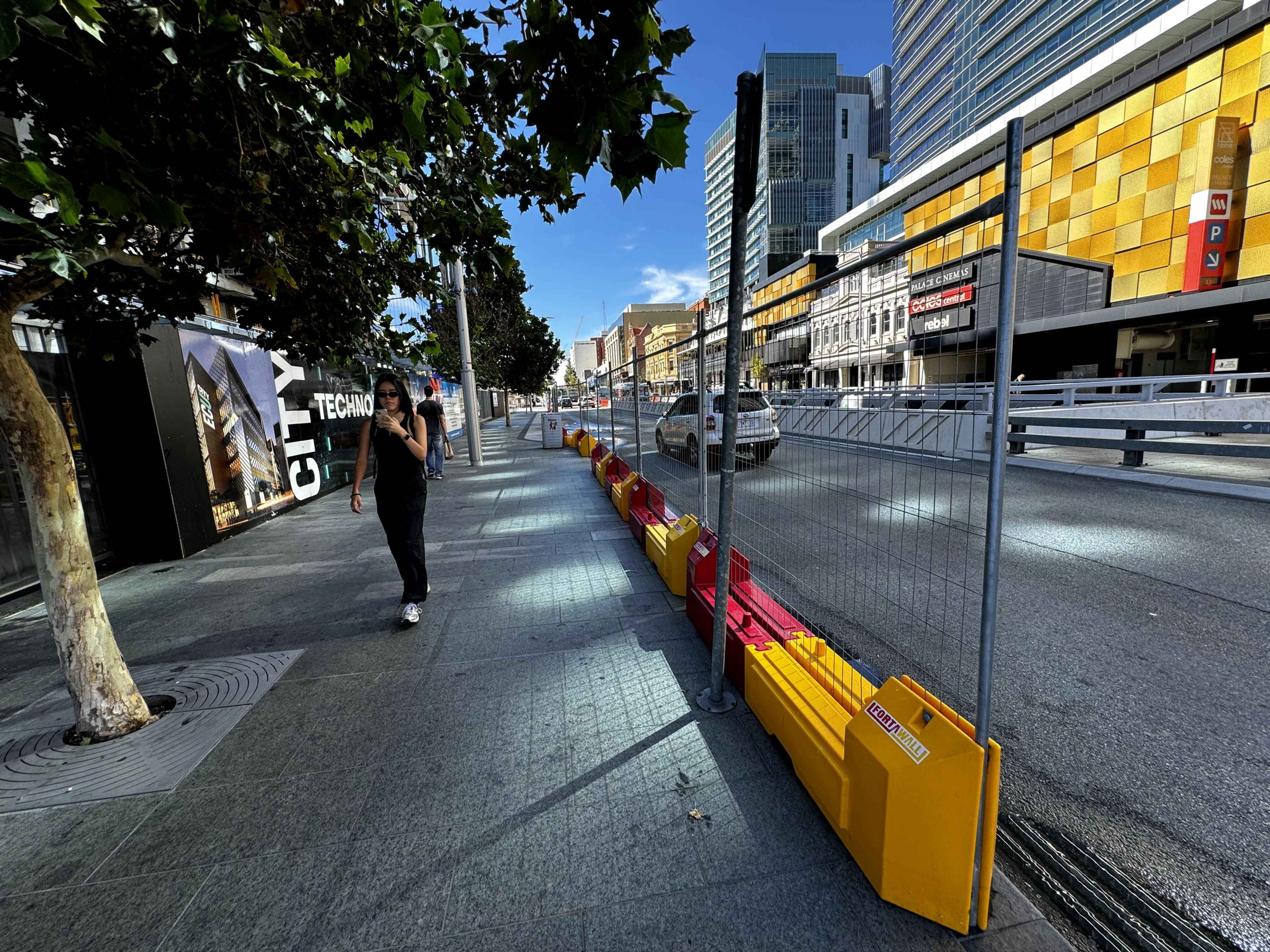 Gallery: City street view with barriers, buildings, and pedestrians. A woman walks on the sidewalk past construction hoarding. Modern architecture.