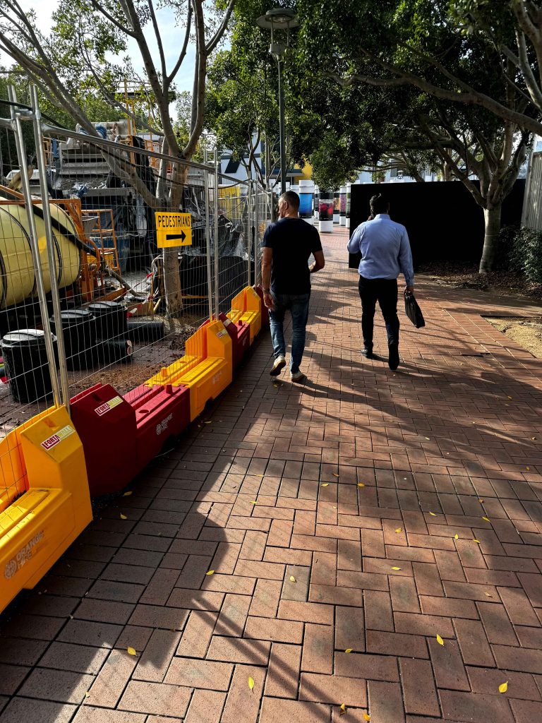 Gallery: Two men walk on a brick path alongside construction barriers and a Pedestrians sign, with trees overhead casting shadows.