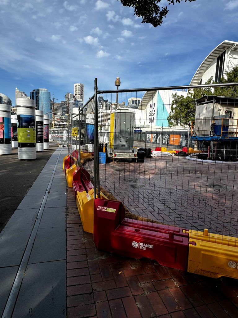 Gallery view of a construction site with barriers, a portable toilet, and the Museum of Contemporary Art in the background, Sydney, Australia.