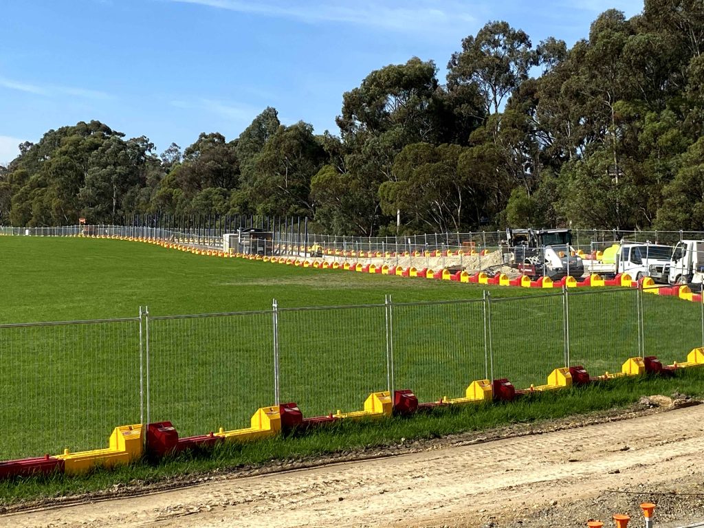 Gallery include: Fenced green field with trees. Red and yellow barriers line the fence. Construction vehicles are visible in the background.