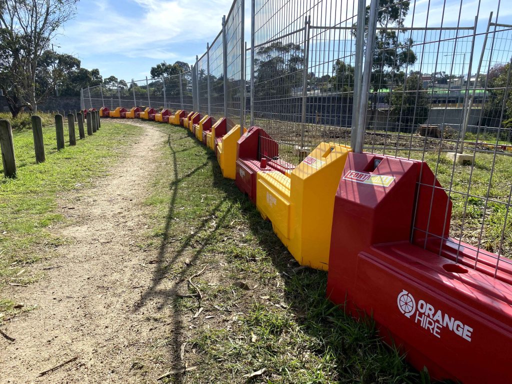 Gallery Include: A dirt path winds between wooden posts and red & yellow barriers, next to a chain link fence. Grass and trees are visible.