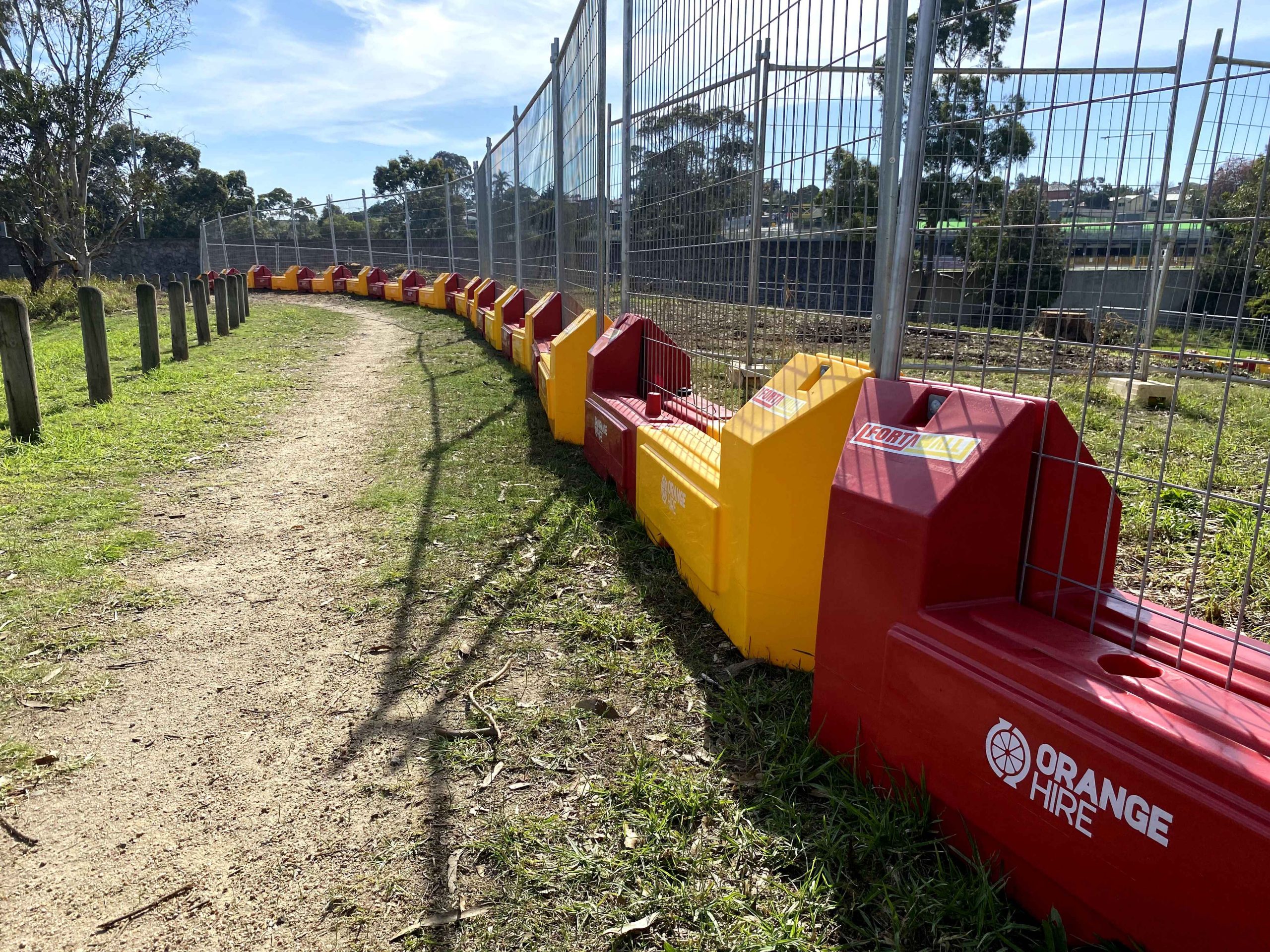 Gallery Include: A dirt path winds between wooden posts and red & yellow barriers, next to a chain link fence. Grass and trees are visible.