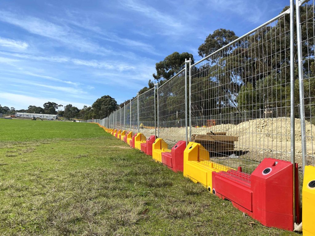 Gallery: A long chain-link fence with red and yellow plastic barriers runs along a grassy field, with trees and a building in the background.
