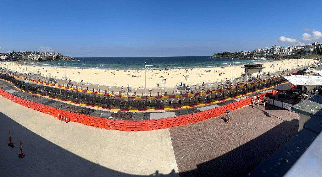 Gallery: Panoramic view of a crowded Bondi Beach, Sydney, Australia, with a lifeguard tower, buildings, and a barricade in the foreground.