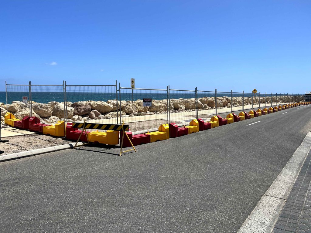 Gallery: Coastal road with rock barrier, wire fence, and red/yellow safety blocks. Ocean view under a clear blue sky. Road stretches into the distance.