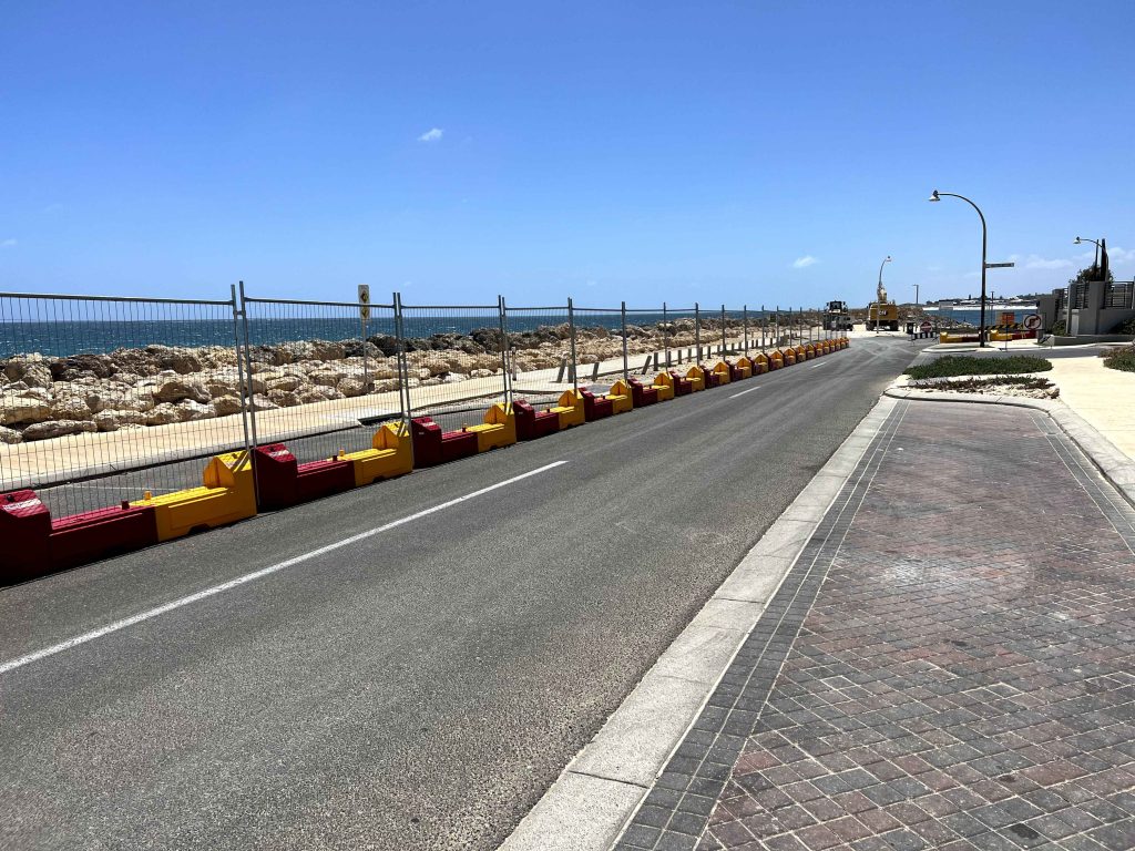 Gallery: Road with barriers along the coast. A fence separates the road from the rocky beach. Clear blue sky above the scene.