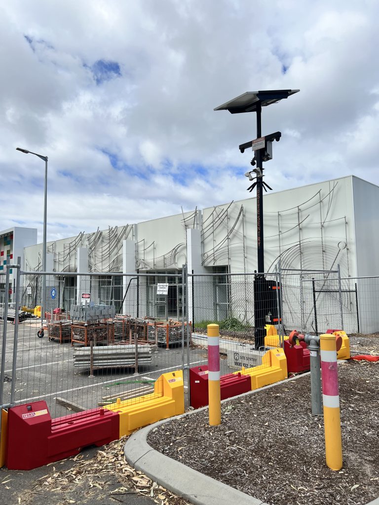 Gallery: Construction site with a solar-powered camera pole behind a safety barrier. Building with artistic metal facade visible in the background.