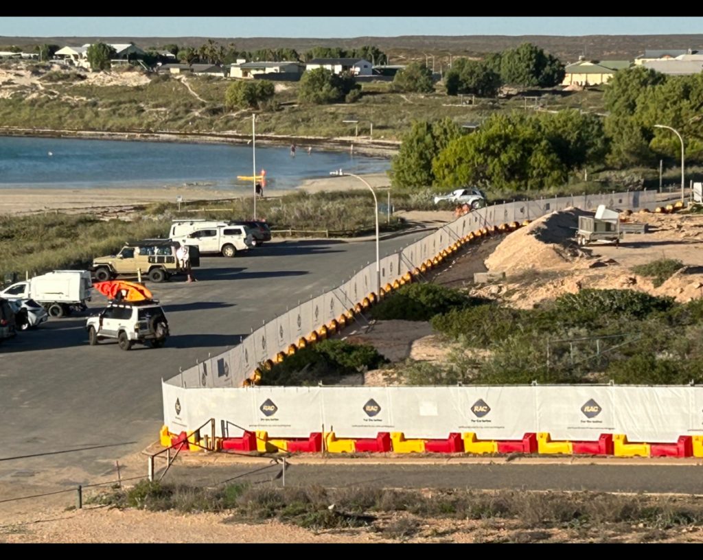Gallery image: Coastal scene with parked 4x4 vehicles near a beach and bay. Construction fencing is visible in the foreground, with houses in the distance.