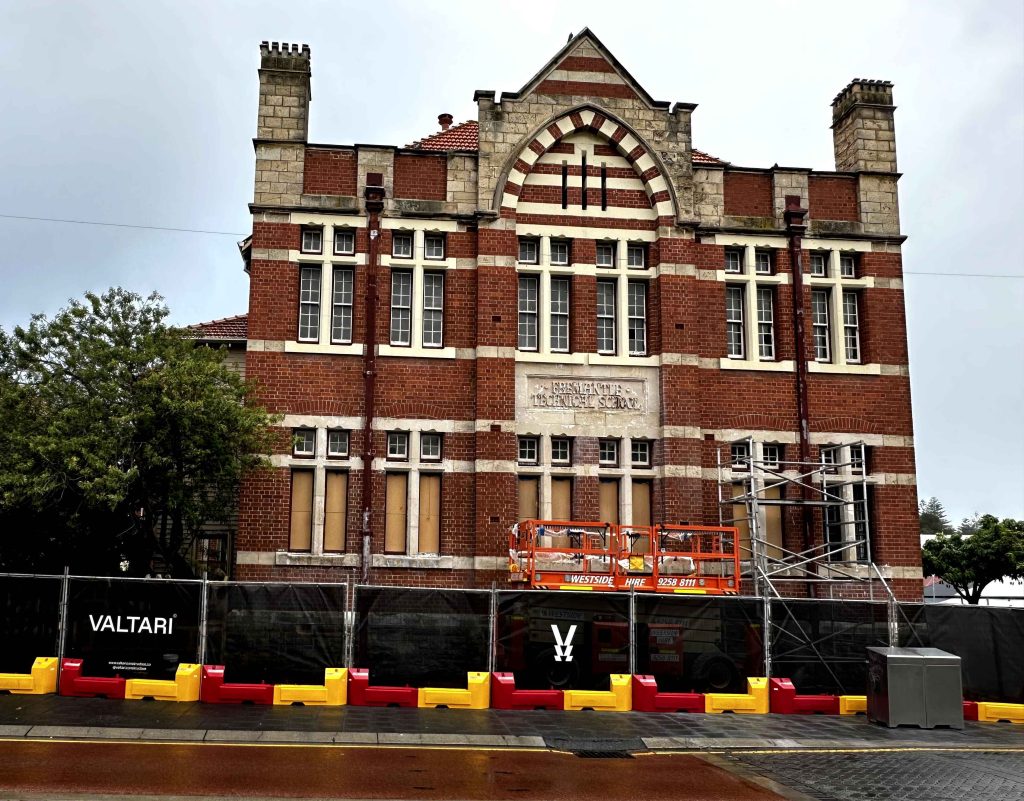 Gallery: Fremantle Technical School, a brick building, is under construction. It features scaffolding, boarded windows, and a safety fence.