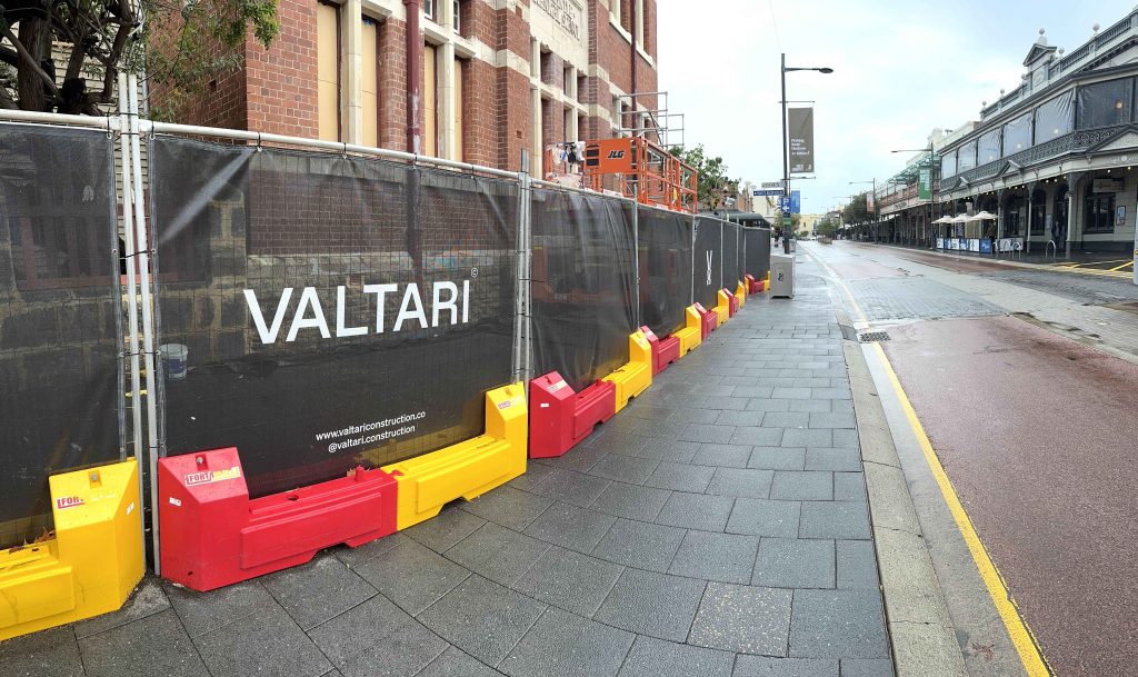 Gallery: Valtari construction fence on a city street. Red and yellow barriers line the sidewalk. Building under construction behind the fence.