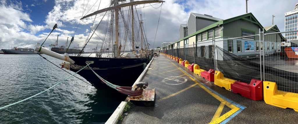 Gallery: Tall ship Leeuwin II docked at a pier. The pier has yellow and red barriers. Buildings are in the background. Cloudy sky above.