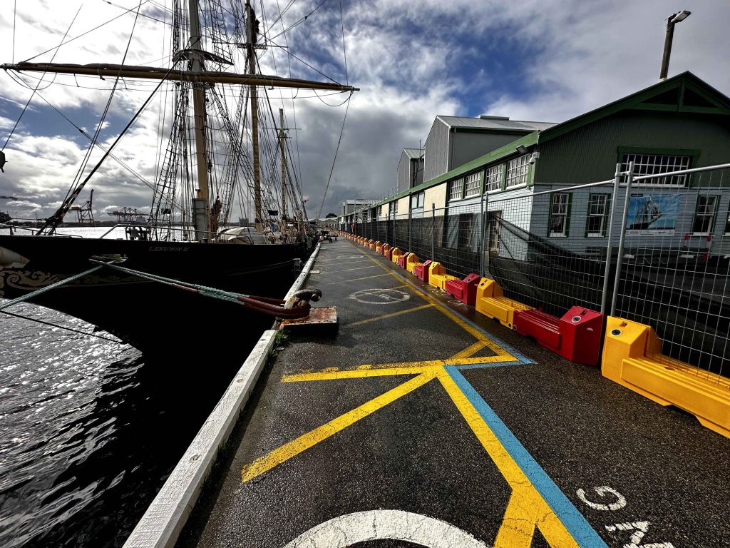Gallery image: Tall ship docked along a pier with yellow and blue markings. Buildings are visible in the background under a cloudy sky.