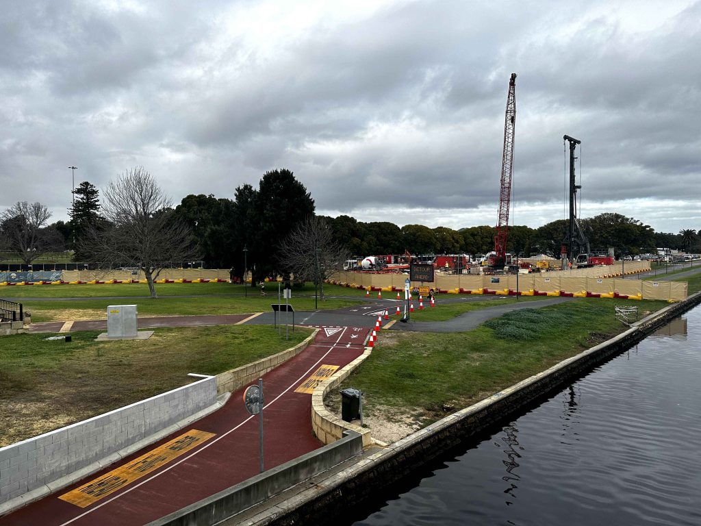Gallery: Construction site with cranes and equipment near a park with a red bike path under a cloudy sky. Water visible.