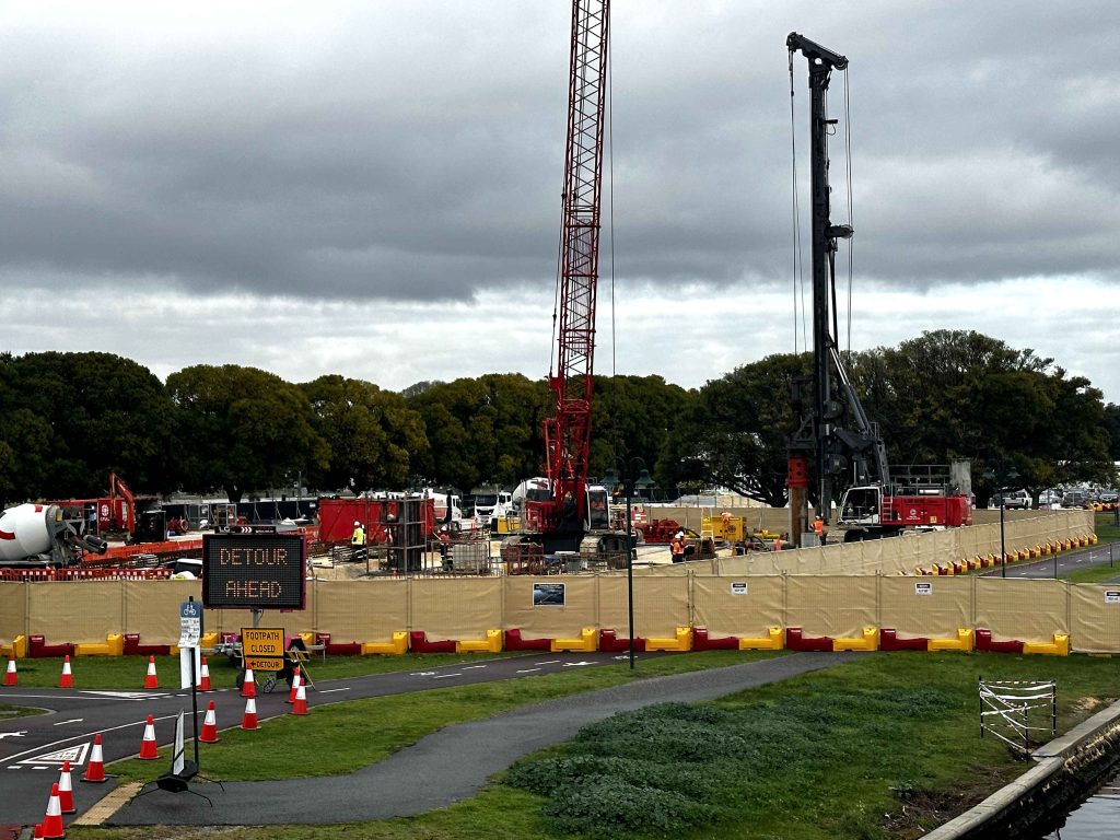 Gallery: Construction site with cranes, heavy machinery, and detour signs. Workers present, surrounded by barriers and safety cones under a cloudy sky.