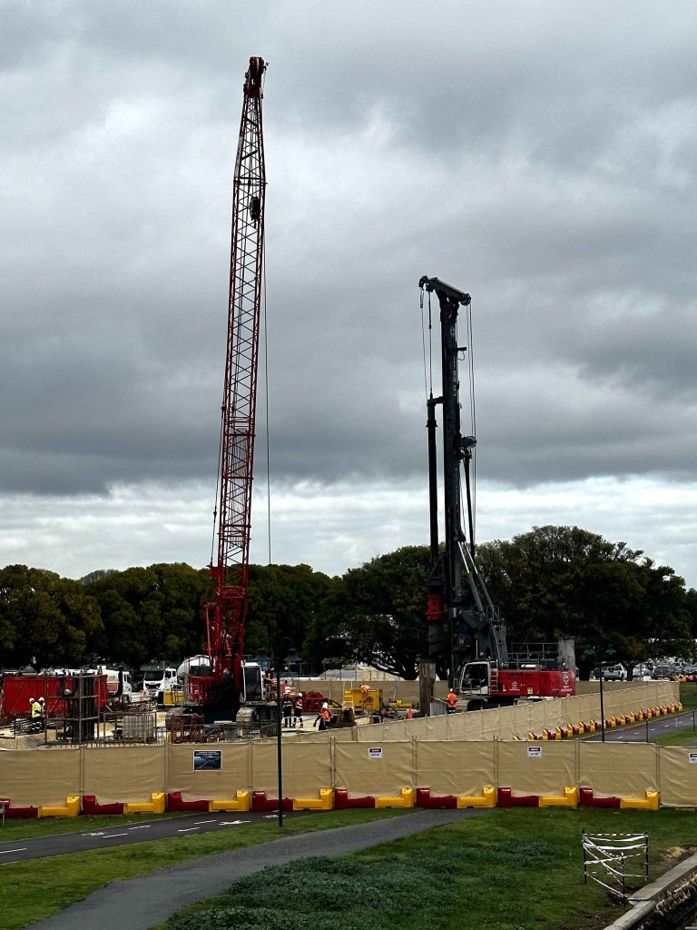 Gallery image: Construction site with a tall red crane and a drilling rig against a cloudy sky. Workers present, safety barriers in place.