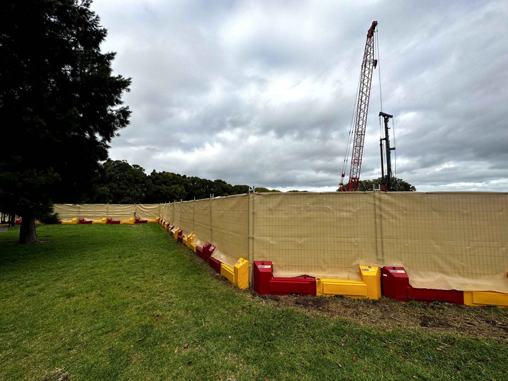 Gallery include: Construction site with a tall red crane visible over a beige fence with red and yellow supports, on a cloudy day.