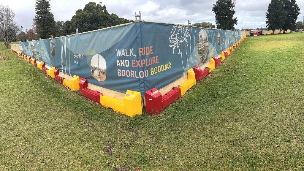 Gallery: Construction fence in a park with Walk, Ride and Explore Boorloo Boodjar banner. Red and yellow barriers line the base.