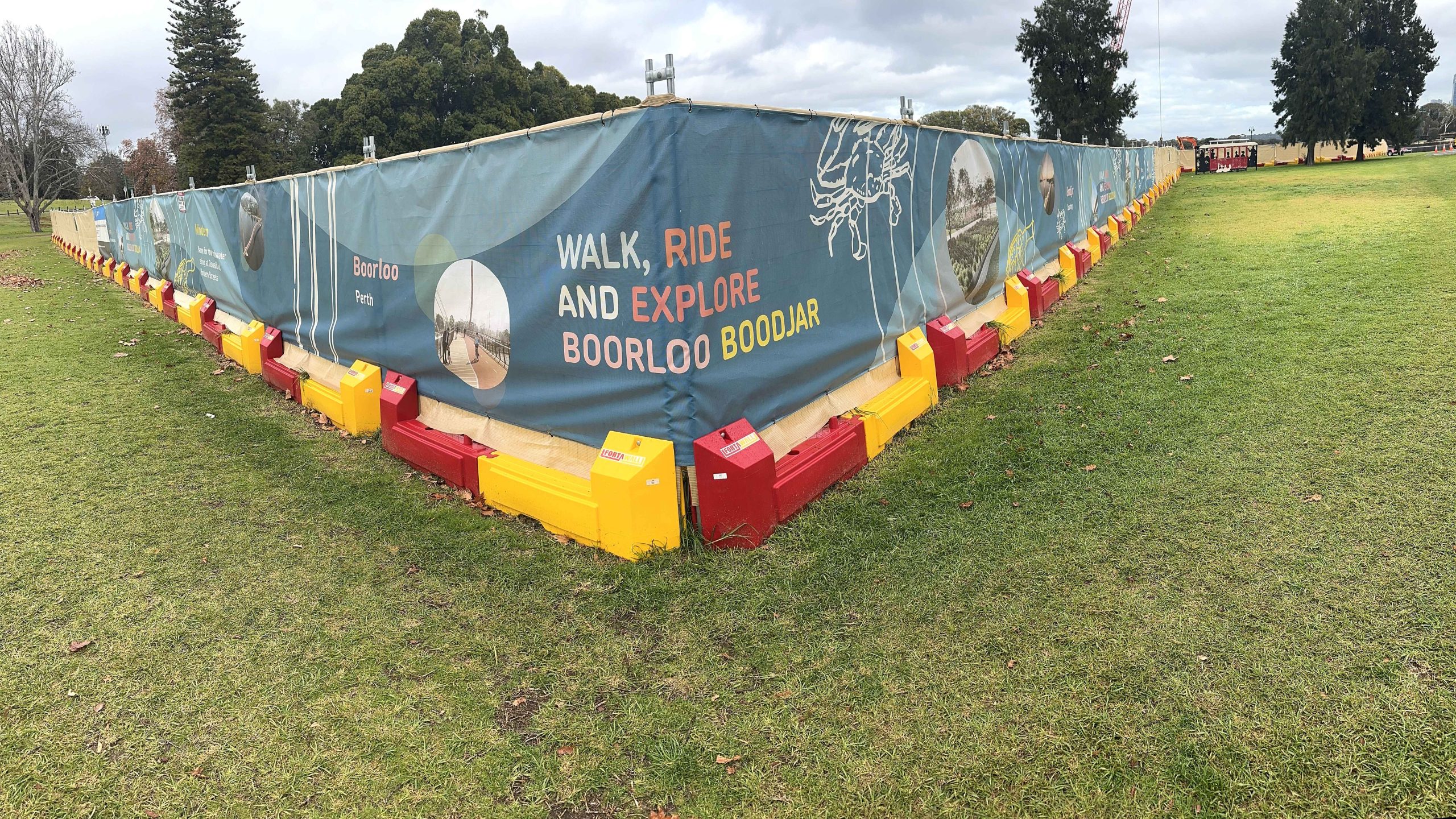 Gallery: Construction fence in a park with Walk, Ride and Explore Boorloo Boodjar banner. Red and yellow barriers line the base.
