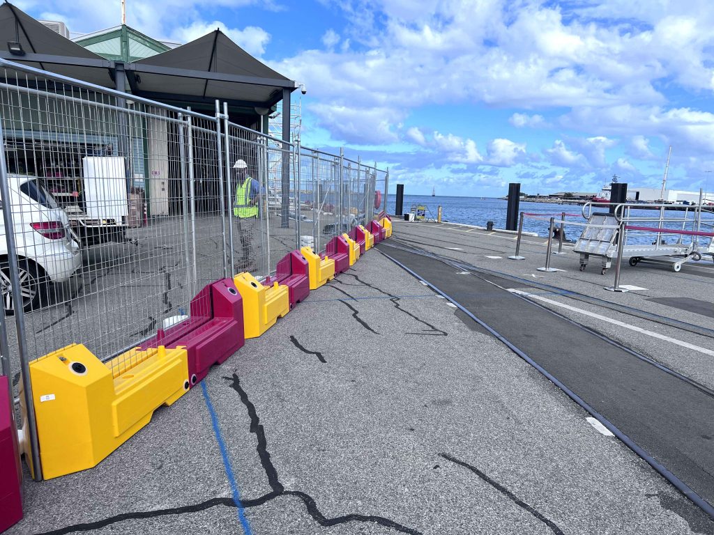 Gallery image: Construction fencing with yellow and red barriers lines a dockside area. A worker stands behind the fence, with the ocean visible in the background.