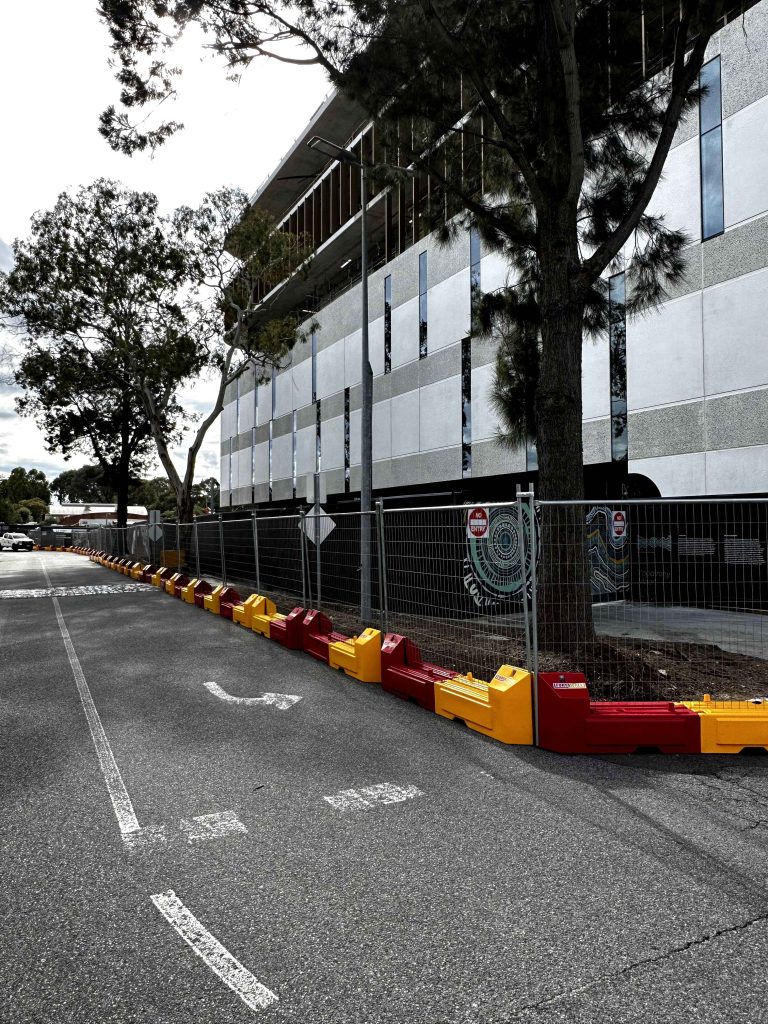 Gallery: Street view with red and yellow barriers lining a fenced area near a modern building. Trees add a natural element to the urban scene.