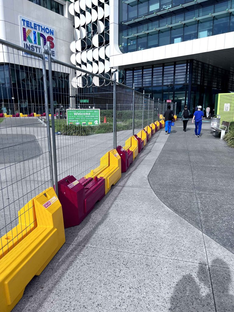 Telethon Kids Institute building with construction barriers. People walk on a sidewalk. Gallery image of the exterior, featuring modern architecture and pedestrian access.