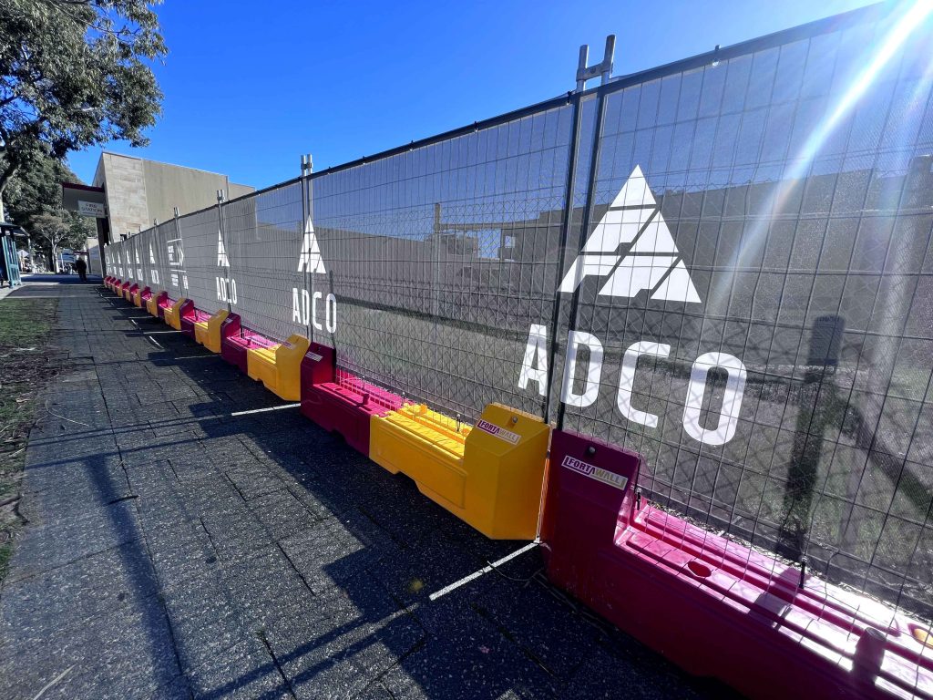 Gallery: Construction site barrier with ADCO logo, featuring a mesh fence, red and yellow FortaWall bases, and a clear blue sky.