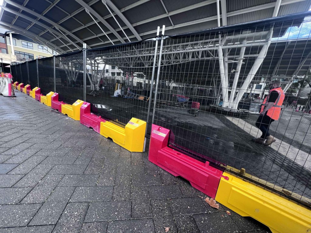 Gallery: Construction site fenced off with pink and yellow barriers. A worker in an orange vest stands nearby. Overhead shelter structure visible.