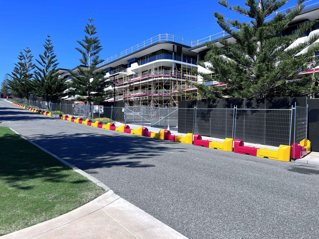 Gallery: Building under construction behind a safety fence on a sunny day. Tall pine trees line the street. Scaffolding visible.