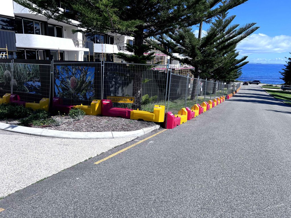Gallery: Street view with barriers lining a construction site fence. Trees and a building are visible, with the ocean in the background under a blue sky.