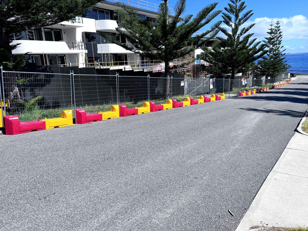 Gallery image: Roadside view of red and yellow barriers lining a construction site with a building under renovation, framed by tall pine trees and the ocean.