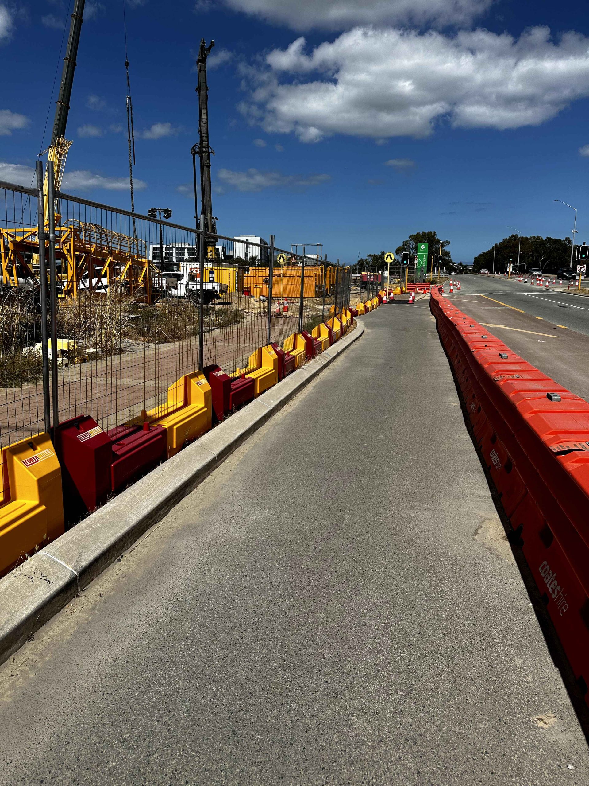 Gallery: Construction site with cranes behind a fence and yellow barriers. Red barriers line the road, under a blue sky with clouds.
