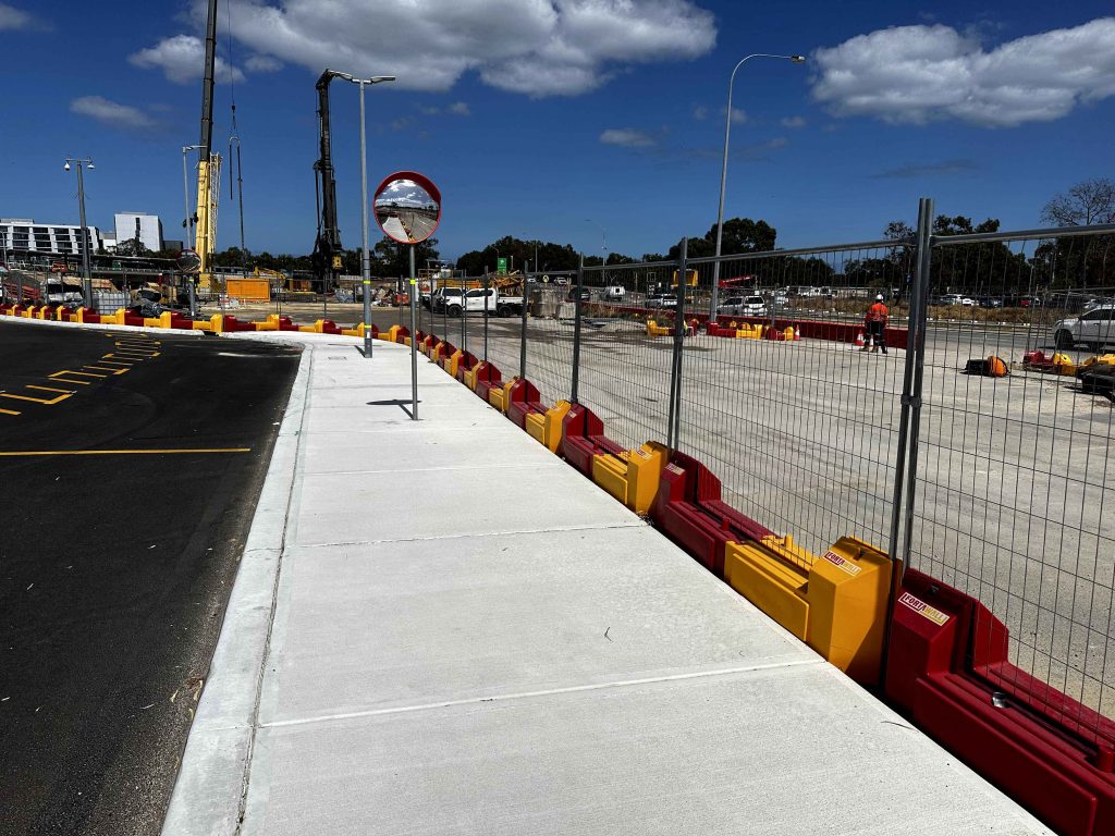 Gallery: Construction site with red and yellow barriers, a sidewalk, and a safety mirror. Cranes and construction equipment are visible in the background.