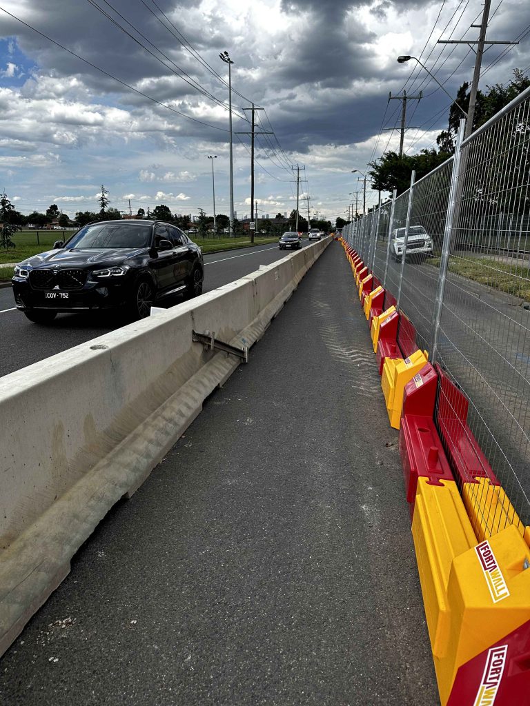 Gallery: Roadside view with concrete barriers and FortaWall fencing separating traffic. Cars visible on the road under a cloudy sky.
